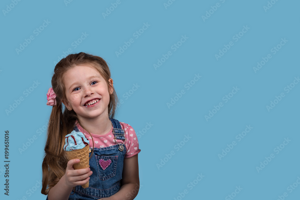 The little girl cheerfully eats ice cream in wafer gunny on blue background in studio. The little girl cheerfully eats ice cream in wafer gunny on blue background in studio.