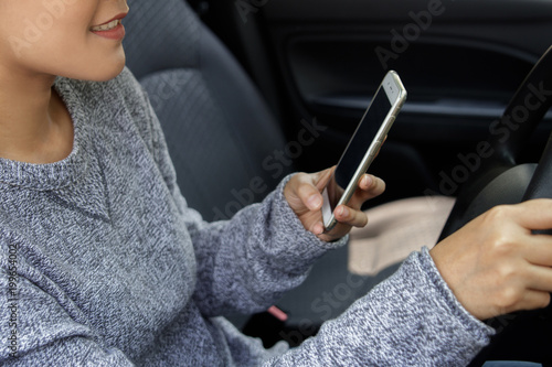 Woman in casual wear driving a car, using smartphone and smiling. Risky driving. Woman sitting in car with cell phone.