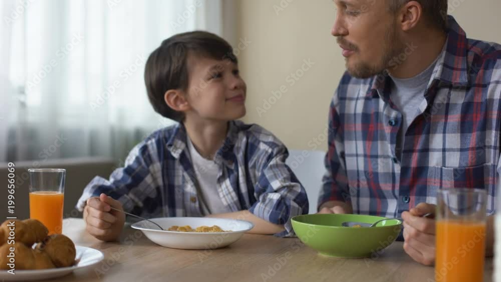 Pleased son and father eating tasty cornflakes at breakfast, morning tradition Pleased son and father eating tasty cornflakes at breakfast, morning tradition