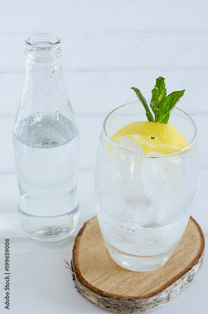 Sparkling water in glass cup with lemon and mint branch on white wooden background.