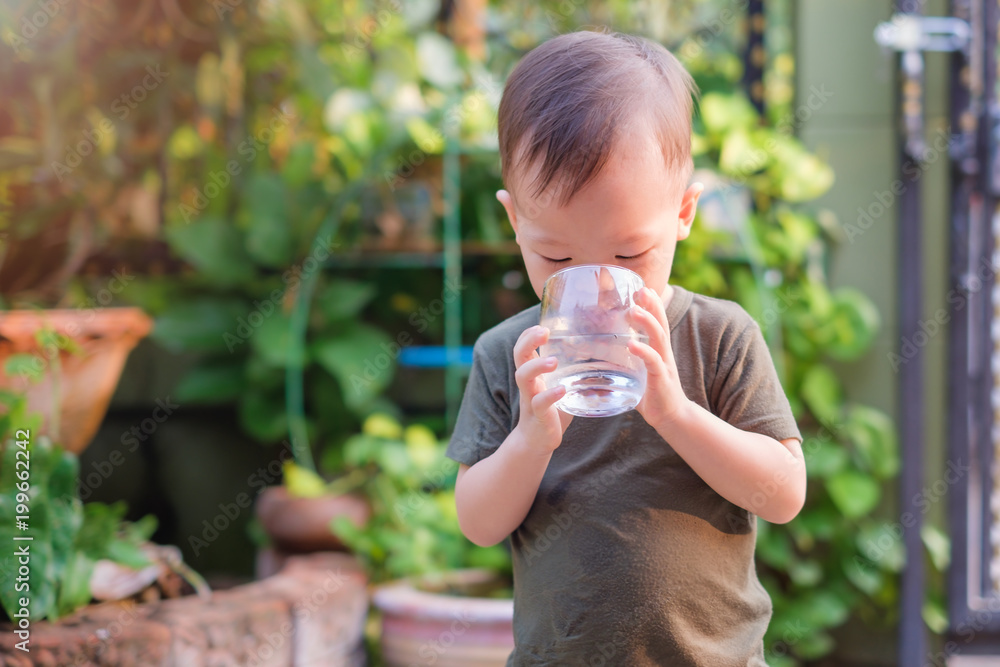 Cute thirsty little Asian 18 months / 1 year old toddler boy child holding and drinking glass of water by himself against green background near home garden, best beverages for child's health concept Cute thirsty little Asian 18 months / 1 year old toddler boy child holding and drinking glass of water by himself against green background near home garden, best beverages for child's health concept