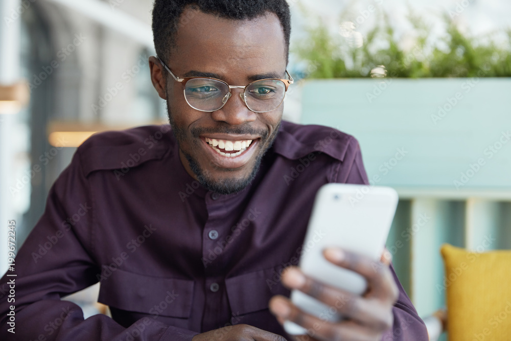 Cheerful young businessman in round spectacles and formal wear, checks newsfeed on modern smart phone, connected to wireless internet, has happy look as recieves positive news from colleagues Cheerful young businessman in round spectacles and formal wear, checks newsfeed on modern smart phone, connected to wireless internet, has happy look as recieves positive news from colleagues
