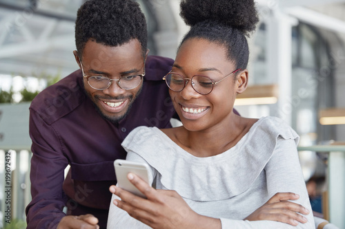 Happy African female in spectacles types message on modern smart phone while her boyfriend stands near her and looks at screen, have broad smile and white teeth. Dark young skinned woman and man