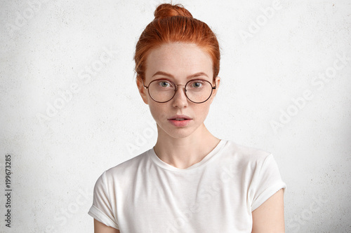 Photo of serious clever redhead female student with ginger hair knot, wears round spectacles, comes on lecture at university, isolated over white concrete background. Confident ginger woman in eyewear