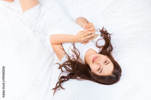 Young happy asian woman laying on her bed using her smartphone to connect to her friends on social media