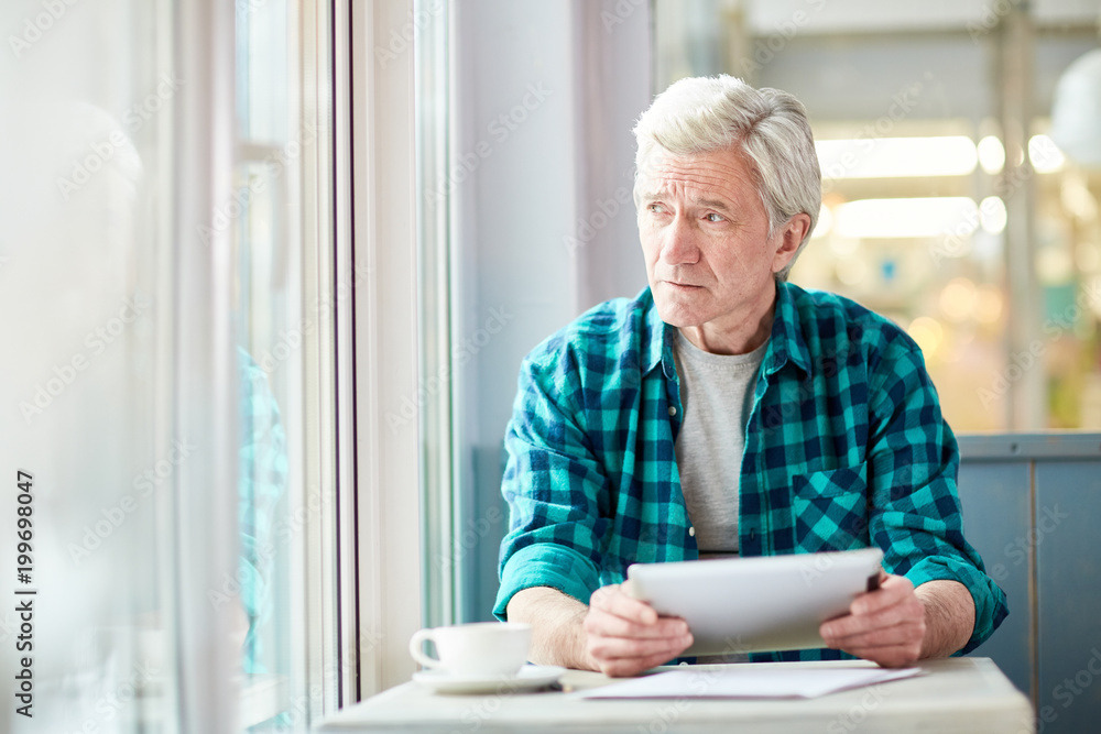 Pensive senior man with tablet looking through window of cafe while thinking of working points Pensive senior man with tablet looking through window of cafe while thinking of working points