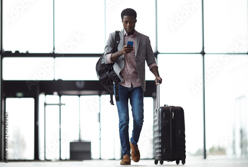 Young mobile traveler with smartphone, backpack and suitcase walking along airport and messaging or looking through online information