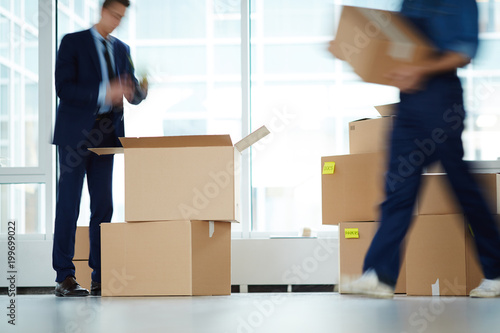 Businessman standing over open box by window of office center while relocation service worker carrying packages photo