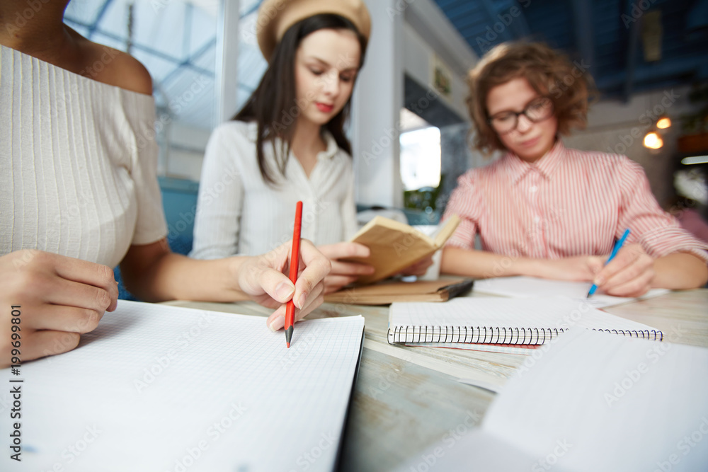 Girl with red pencil going to draw something on page of notepad with her busy groupmates on background Girl with red pencil going to draw something on page of notepad with her busy groupmates on background