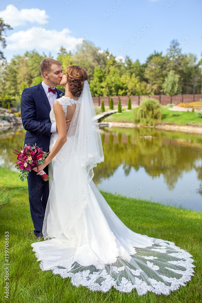 happy groom and bride kissing while standing on green grass near pond at summer day
