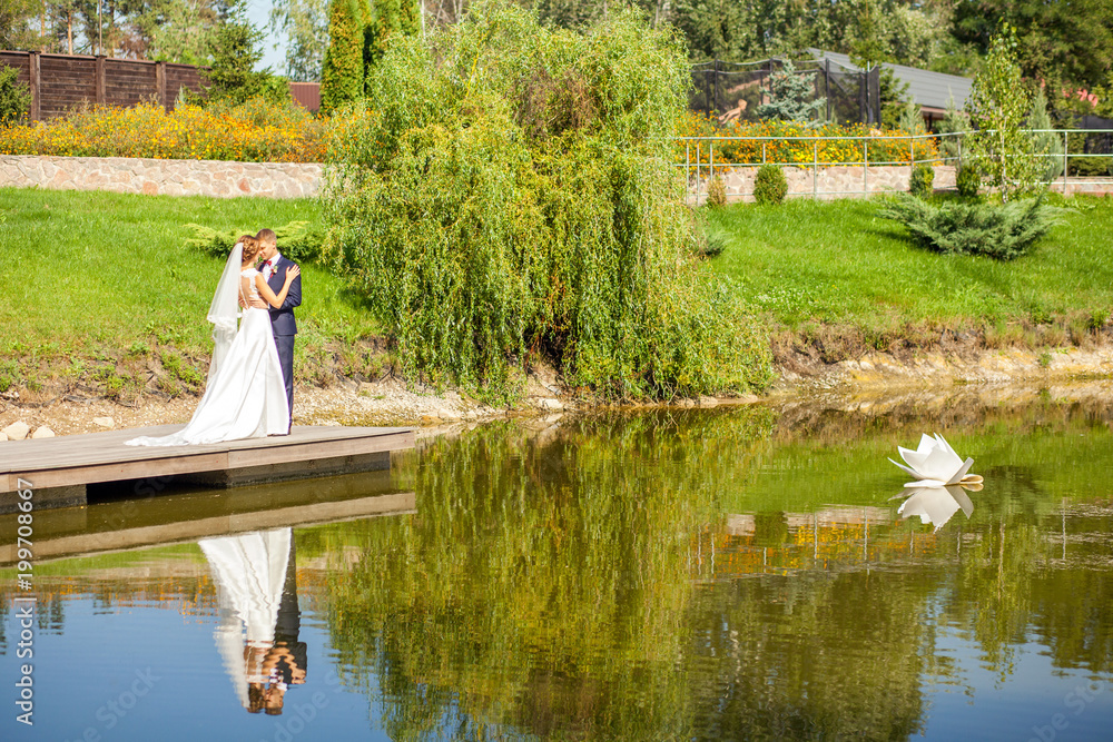 newlyweds hugging while standing on wooden pier near lake at sunny summer day newlyweds hugging while standing on wooden pier near lake at sunny summer day