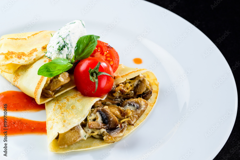 Homemade tasty pancakes (blinis) served with mushrooms and tomatoes in the white plate on dark background Homemade tasty pancakes (blinis) served with mushrooms and tomatoes in the white plate on dark background