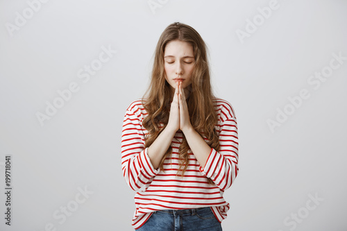 Praying together for peace. Portrait of worried upset woman holding hands in pray and closing eyes while talking with god, wishing and hoping to solve issue, begging for forgiveness over gray wall