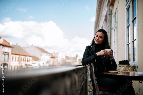 Attractive girl with long hair holding a cup on a balcony in city.