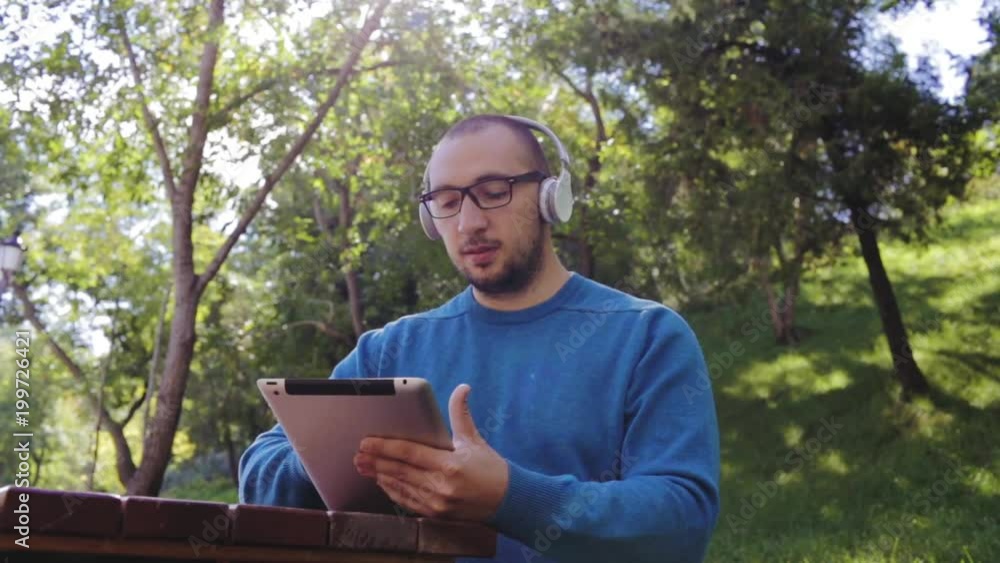 Technology, lifestyle and people concept - smiling young man or teenager with tablet and headphones listening to music outdoors. Cheerful teenage boy listening to music with headphones. Technology, lifestyle and people concept - smiling young man or teenager with tablet and headphones listening to music outdoors. Cheerful teenage boy listening to music with headphones.