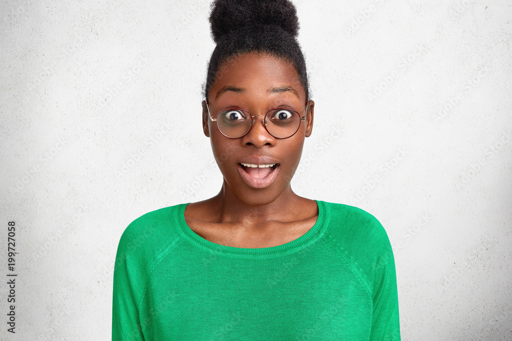 Photo of amazed dark skinned female looks with bated breath and unexpected expression, wears casual green sweater and round glasses, recieves surprise from husband. Emotional African young woman Photo of amazed dark skinned female looks with bated breath and unexpected expression, wears casual green sweater and round glasses, recieves surprise from husband. Emotional African young woman