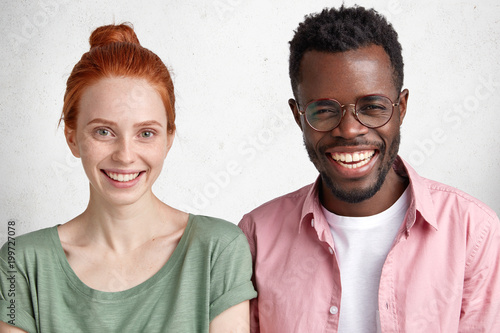 Interracial friendship concept. Beautiful freckled woman with healthy skin and dark skinned overjoyed male wears spectacles, giggles joyfully, being in good mood after successful passed exam