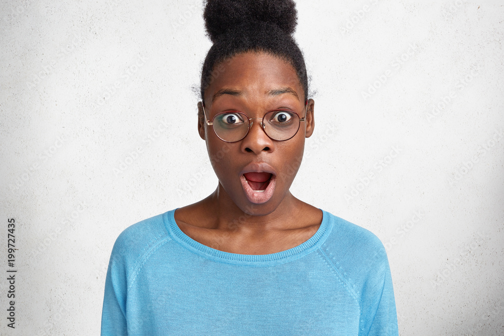 Shocked middle aged dark skinned African American female has stunned expression, gets much work and little time for preparation, wears casual blue sweater, isolated over white concrete wall.