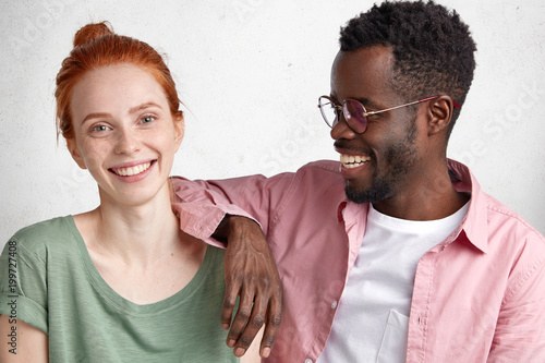 Cheerful young European beautiful woman with ginger hair knot, dressed casually ,enjoys nice time in company of handsome African American male who leans at her shoulder, smile with enjoyment