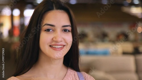 Portrait of a stylish young woman with long loose hair standing and smiling open heartedly in a modern shopping mall. She feels cheerful photo