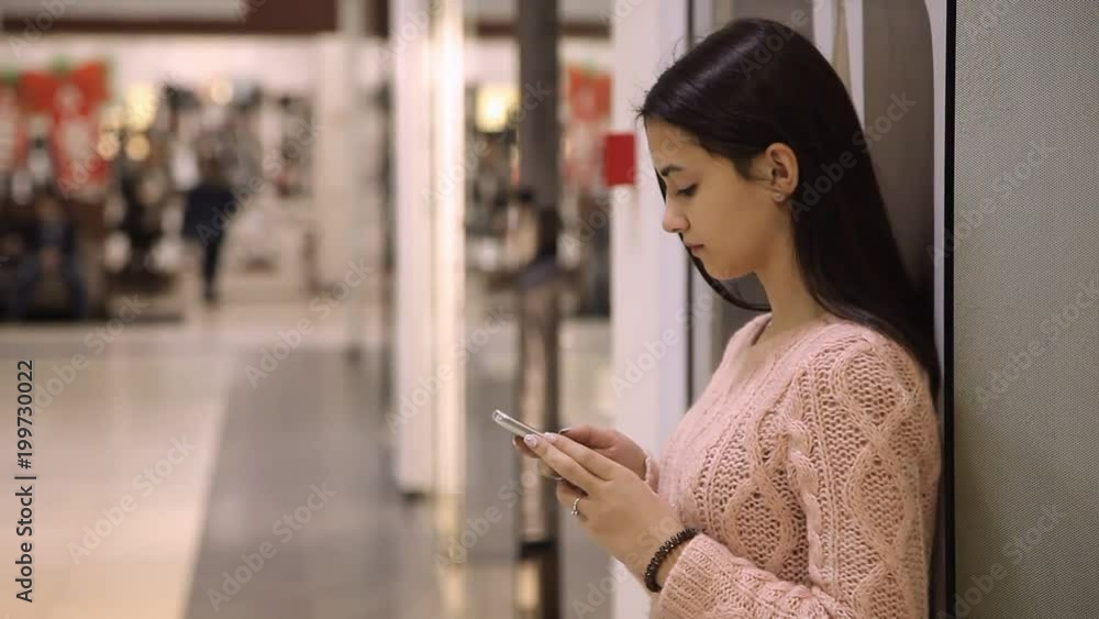 Profile of ah elegant young woman touching the screen of her smartphone and seeking some business information while standing in a shopping mall Profile of ah elegant young woman touching the screen of her smartphone and seeking some business information while standing in a shopping mall