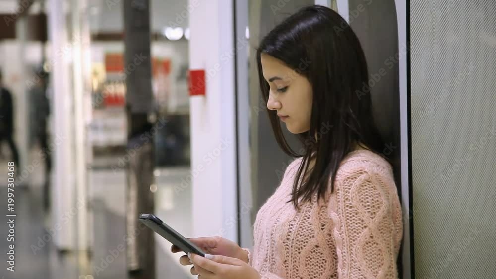 Profile of a charming young woman touching the screen of her tablet and seeking some product while standing in a glassy shopping mall Profile of a charming young woman touching the screen of her tablet and seeking some product while standing in a glassy shopping mall