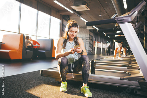 Beautiful young Caucasian girl sportswoman sitting, resting after training on treadmill against the backdrop of gym in sunny weather. Listens to music in sick white headphones,in hand holds the phone photo