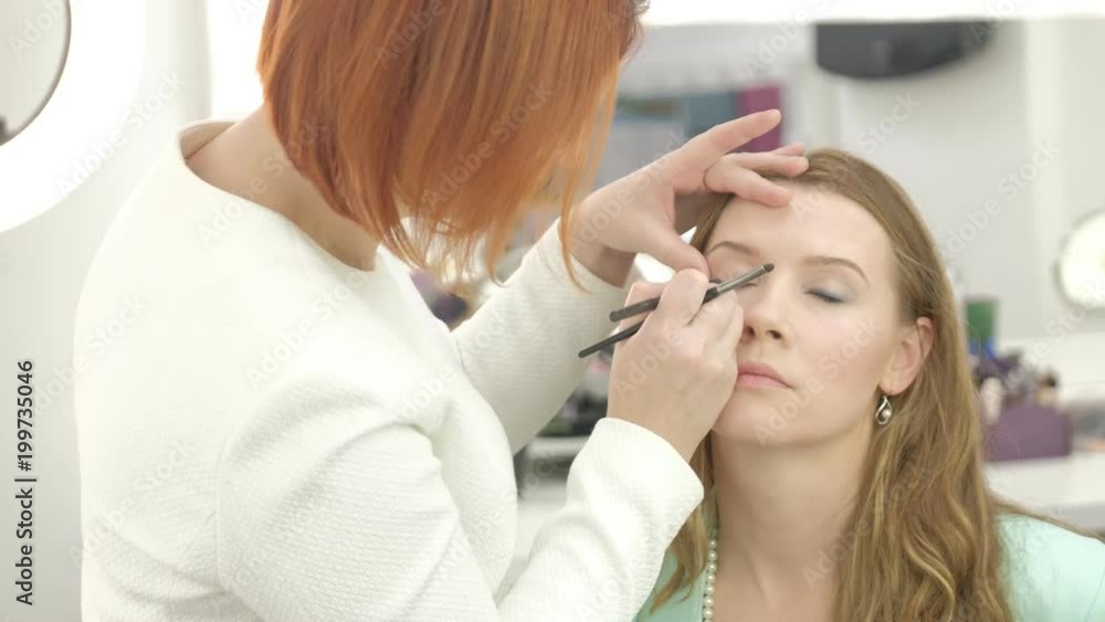Portrait of young beautiful woman and stylist in make up salon brushing eyelashes holding set of colors Portrait of young beautiful woman and stylist in make up salon brushing eyelashes holding set of colors