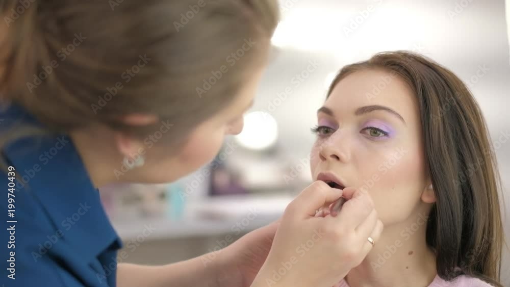 Portrait of young beautiful woman dressed pink sitting in beauty salon and stylist apply lipstick Portrait of young beautiful woman dressed pink sitting in beauty salon and stylist apply lipstick