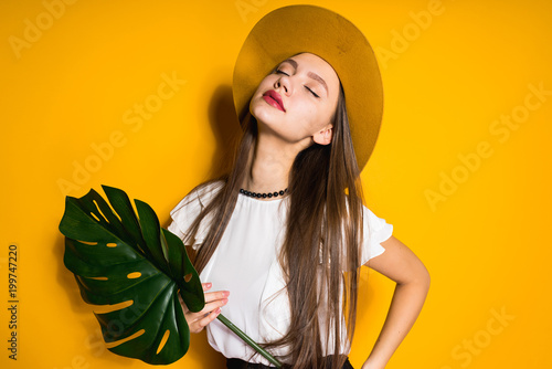 confident long-haired girl model in a fashionable hat posing on a yellow background, holding a green leaf, closing her eyes