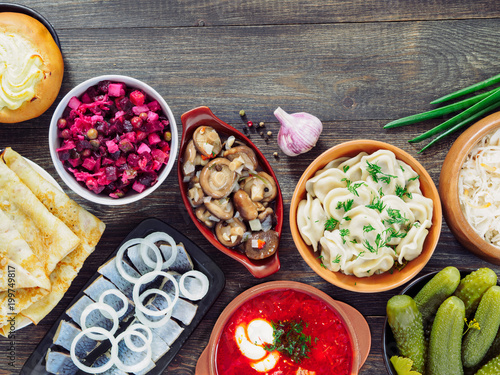 Russian food on wooden background. Assortment dishes of Russian cuisine - borscht, pelmeni, herring, marinated mushrooms, salted cucumbers, vinaigrette, sauerkraut and pancakes. Top view. Copy space.