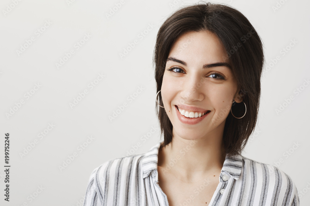 Attractive european shop assistant in stylish earrings and blouse smiling broadly and politely while gazing at camera, wanting to help customer to pick gift for girlfriend, standing over gray wall Attractive european shop assistant in stylish earrings and blouse smiling broadly and politely while gazing at camera, wanting to help customer to pick gift for girlfriend, standing over gray wall