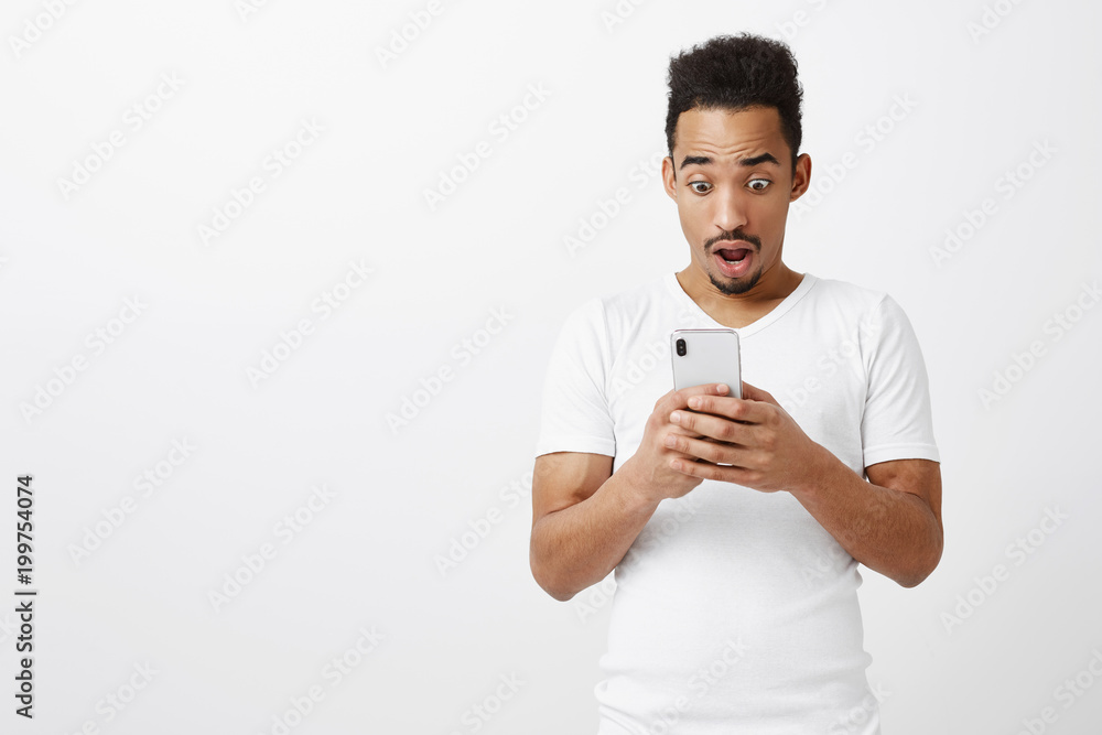 Worried guy checked his bank account, being poor. Stunned and shocked good-looking african man in ordinary outfit, holding smartphone, dropping jaw from reading surprising message over gray wall Worried guy checked his bank account, being poor. Stunned and shocked good-looking african man in ordinary outfit, holding smartphone, dropping jaw from reading surprising message over gray wall