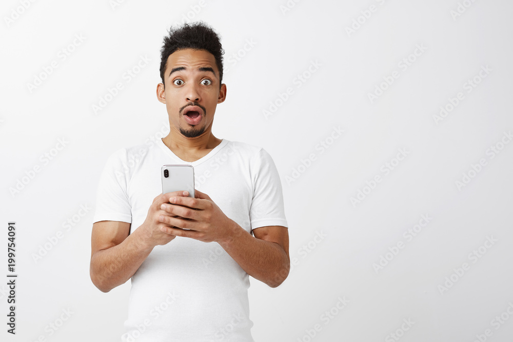 Hair stands from shock. Good-looking young african-american student with afro hairstyle holding smartphone and dropping jaw from surprise or amazement, being over-emotional over gray background Hair stands from shock. Good-looking young african-american student with afro hairstyle holding smartphone and dropping jaw from surprise or amazement, being over-emotional over gray background