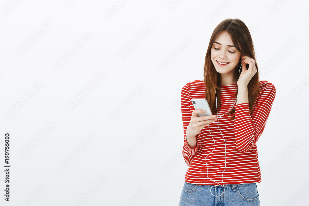 Time to cheer up with music in ears. Portrait of cute tender european girlfriend, putting on earphones and picking song in smartphone with joyful happy smile, standing over gray background Time to cheer up with music in ears. Portrait of cute tender european girlfriend, putting on earphones and picking song in smartphone with joyful happy smile, standing over gray background