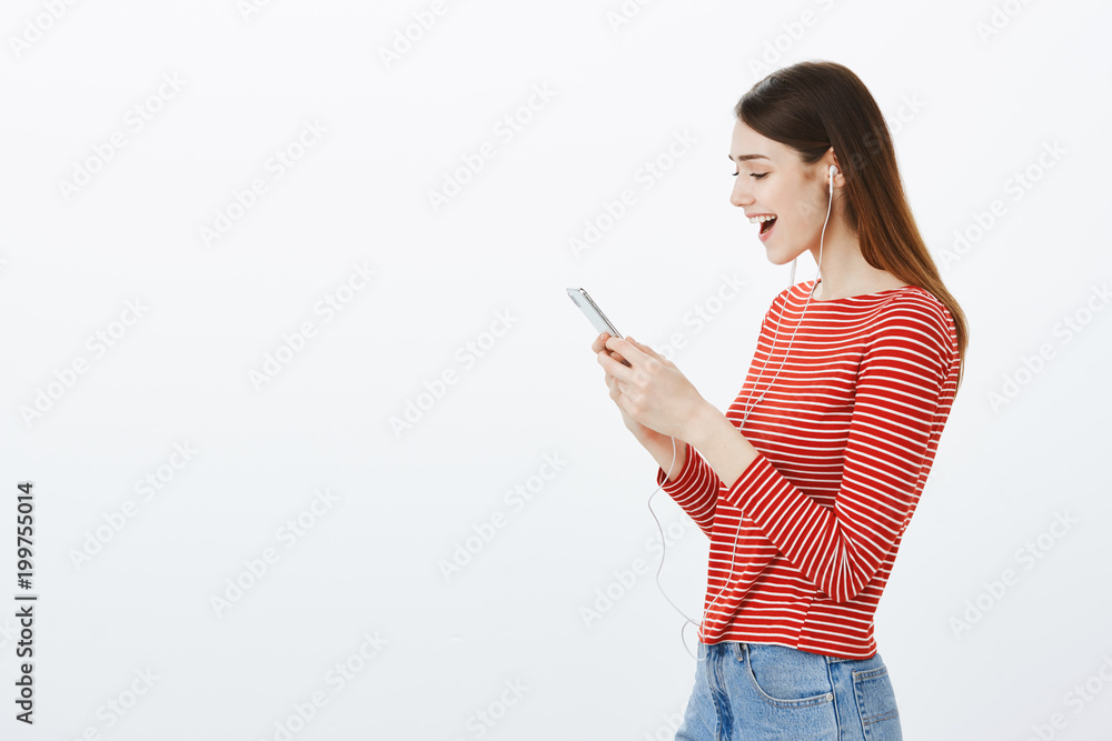 Cheerful bright daughter talking with mom via video chat while studying abroad. Relaxed carefree good-looking girl, standing half-turned, talking with friends using earphones, looking at smartphone Cheerful bright daughter talking with mom via video chat while studying abroad. Relaxed carefree good-looking girl, standing half-turned, talking with friends using earphones, looking at smartphone