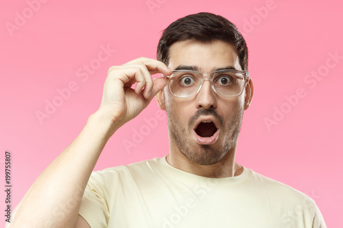 Casual dressed young man in beige t-shirt shouting oh my god with open mouth, surprised by low price and sales, holding transparent glasses, isolated on pink background