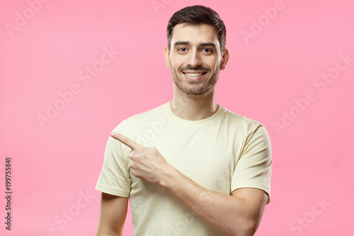 Handsome man in beige t-shirt pointing left with finger isolated on pink background