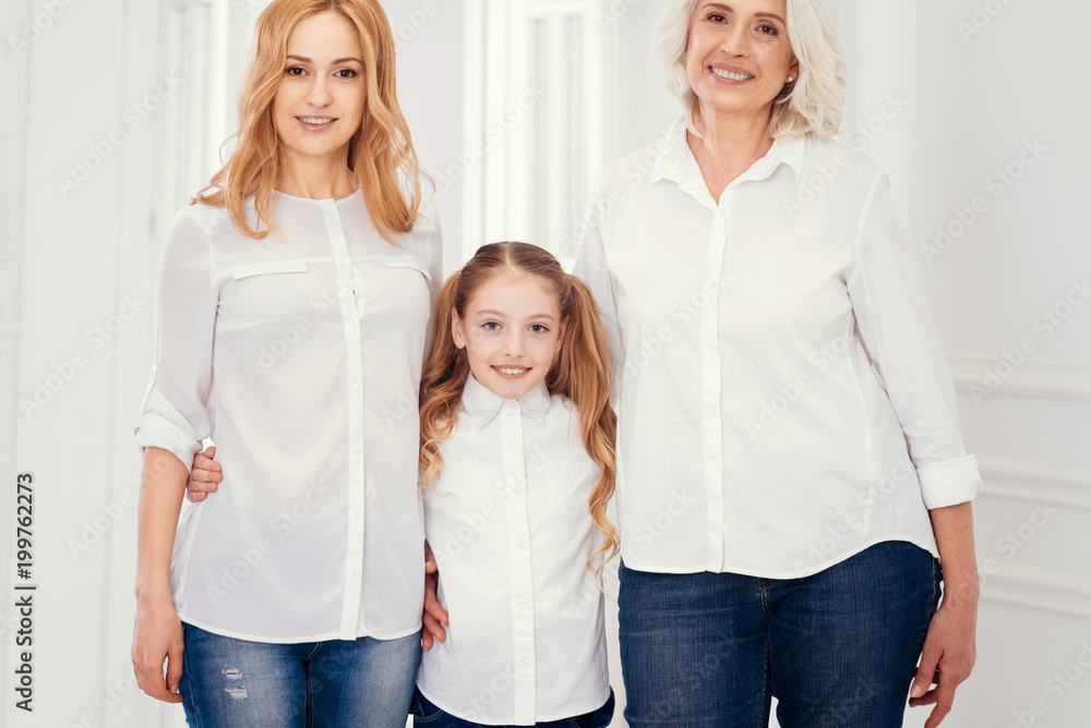 Fototapeta premium Family is forever. Adorable little girl with ponytail standing between her beautiful mother and charming grandmother while all three embracing and smiling for the camera at home.