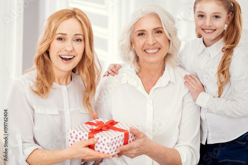 I have no idea what is inside. Amazed lady receiving an unexpected gift box from her loving mother and daughter while all three sitting on a sofa and looking into the camera.