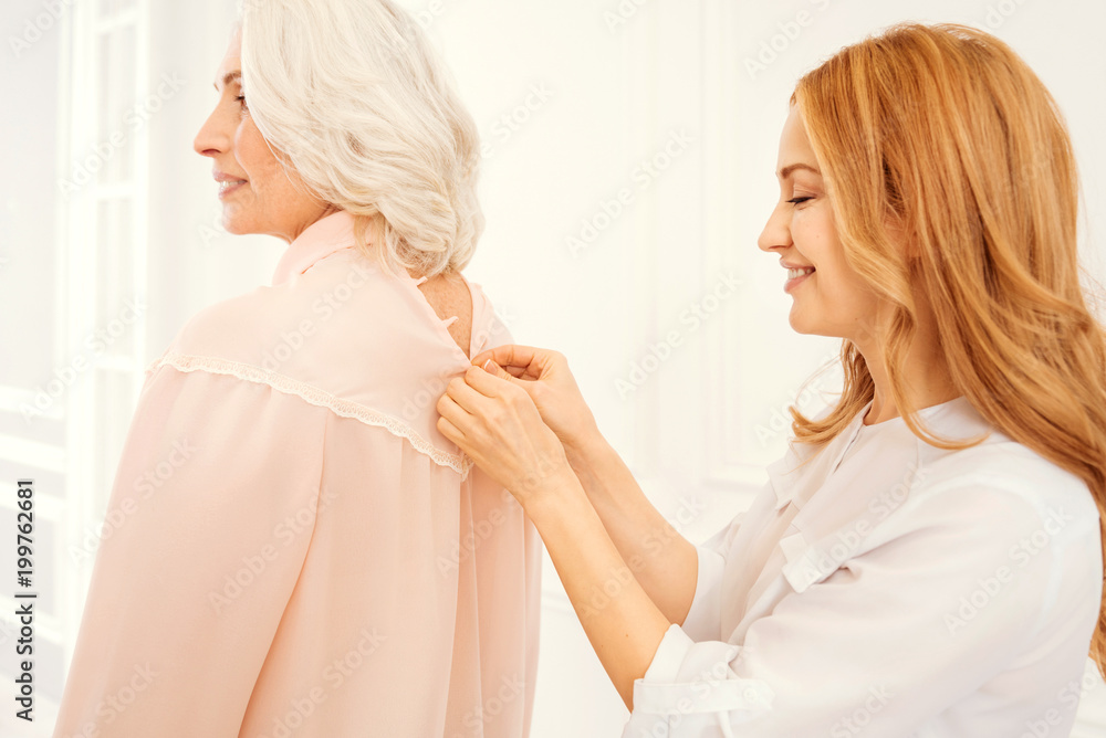 Mother and daughter moment. Side view on a thoughtful adult lady standing behind her beautiful mother and helping her with buttoning a blouse while spending family time together at home. Mother and daughter moment. Side view on a thoughtful adult lady standing behind her beautiful mother and helping her with buttoning a blouse while spending family time together at home.