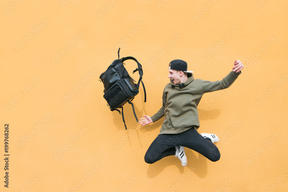 Street young man jumping with a backpack on the background of an orange wall - obrazy, fototapety, plakaty 