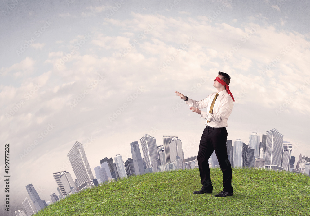 Young blindfolded businessman steps on a a patch of grass with a grey buildings in the background Young blindfolded businessman steps on a a patch of grass with a grey buildings in the background
