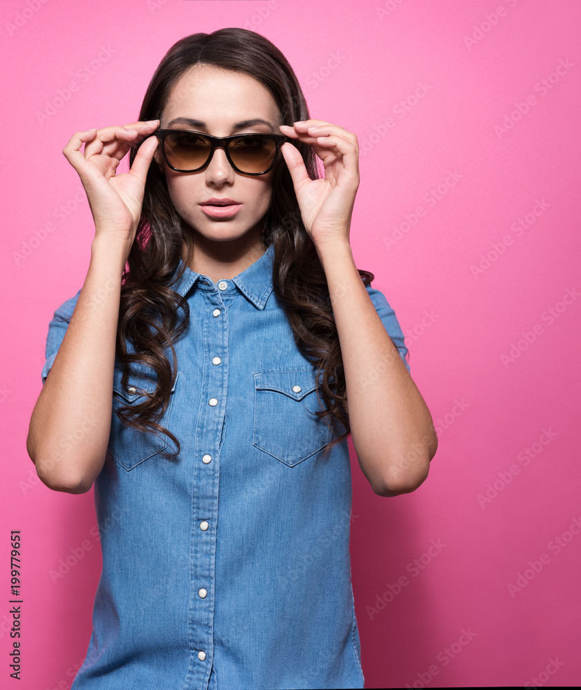 Attractive young woman with a neutral expression holds for sunglasses. Stylish look, blue denim shirt Attractive young woman with a neutral expression holds for sunglasses. Stylish look, blue denim shirt