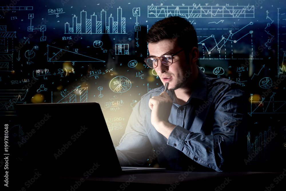Young handsome businessman working late at night in the office with blue calculations in the background Young handsome businessman working late at night in the office with blue calculations in the background