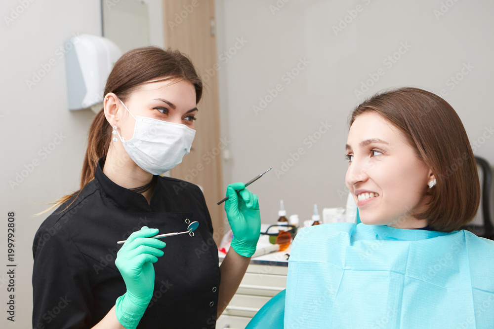 Candid shot of scared anxious woman sitting in dental chair while going through dental examination, looking at female dentist in white mask who is holding metal probe with small angled mirror Candid shot of scared anxious woman sitting in dental chair while going through dental examination, looking at female dentist in white mask who is holding metal probe with small angled mirror