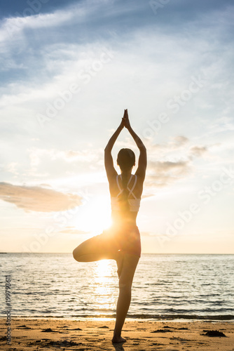 Full length rear view of the silhouette of a woman standing on one leg, while practicing the tree yoga pose on a tranquil beach at sunset during summer vacation in Indonesia