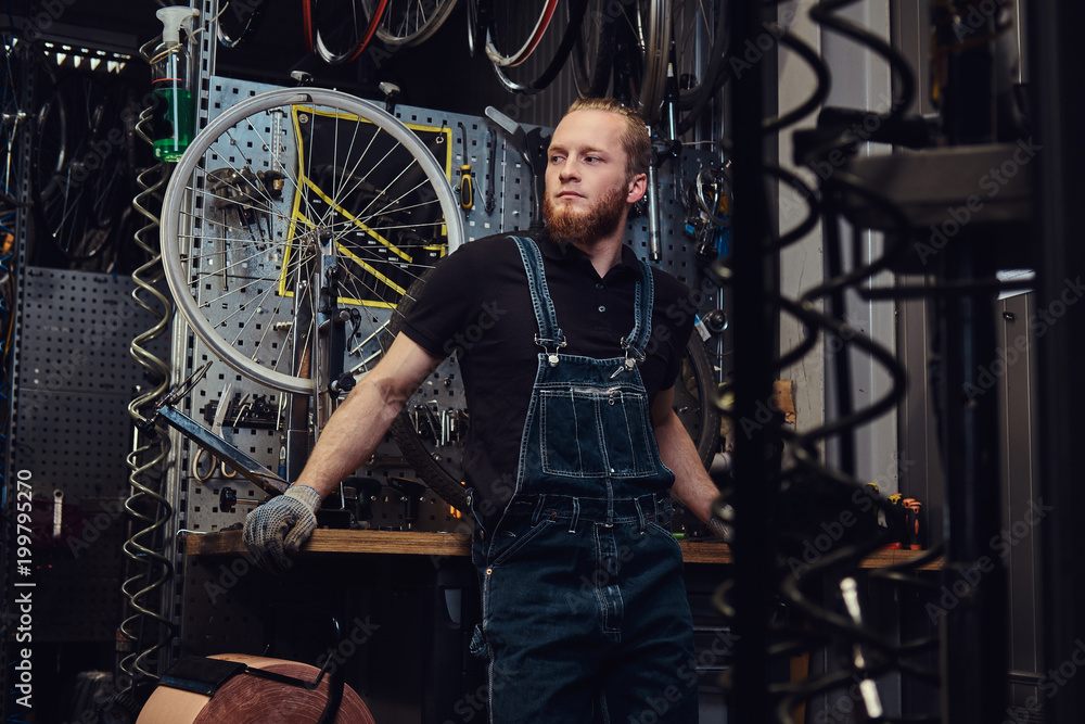Portrait of a handsome redhead male with beard and haircut wearing jeans coverall, standing near bicycle wheel in a workshop against wall tools.