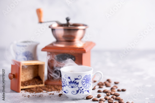 A beautiful coffee composition with a place under your text. Grains of coffee fall out of a vintage coffee grinder. Hot black coffee in a beautiful porcelain cup on the table.