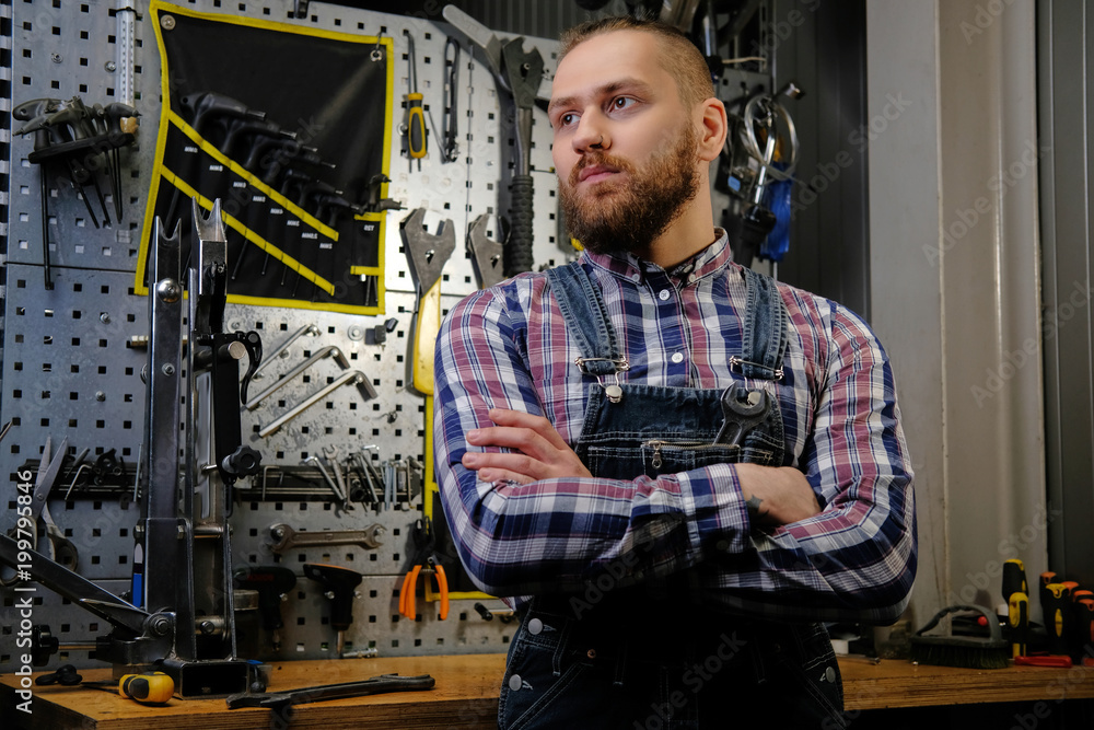 Portrait of a handsome stylish male with beard and haircut wearing a flannel shirt and jeans coverall, standing with crossed arms in a workshop against wall tools.